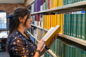 Eleanor Bryan in a library, stood by some shelves, reading a book.